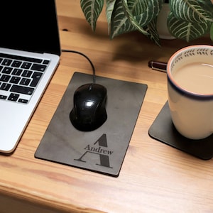 May include: A personalised grey leather mouse mat with the letter "A" and the name "Andrew" engraved on it. A black computer mouse is on the mouse mat.