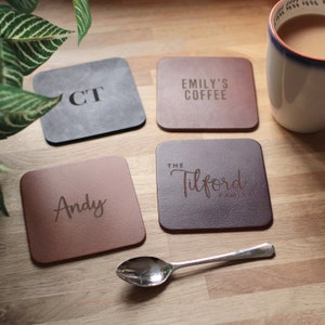 May include: Four square coasters with personalized text. One is gray with "CT", another is brown with "Emily's Coffee", a third is brown with "Andy", and the last is dark brown with "The Tilford Family". A spoon and a mug are also in the image.