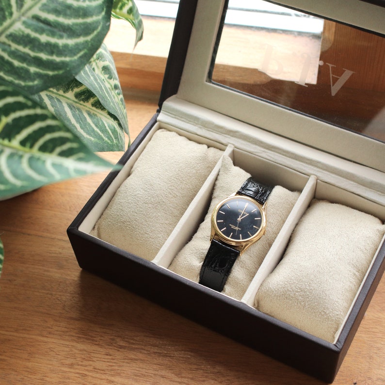 Pu&ograve; includere: Un orologio da polso nero e oro con cinturino in pelle nera &egrave; all'interno di un espositore per orologi marrone. L'orologio ha un quadrante scuro con lancette e indici dorati. La cassa ha un coperchio in vetro e tre scomparti imbottiti beige.