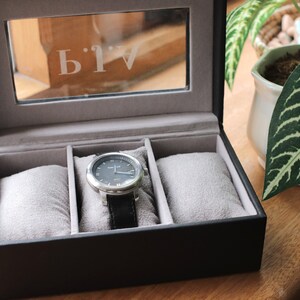 Pu&ograve; includere: Una scatola per orologi nera con un coperchio trasparente, contenente un orologio argentato con un cinturino in pelle nera. L'orologio ha un quadrante grigio scuro. La scatola ha tre scomparti imbottiti grigi. Le lettere "AIV" sono incise sul vetro del coperchio.