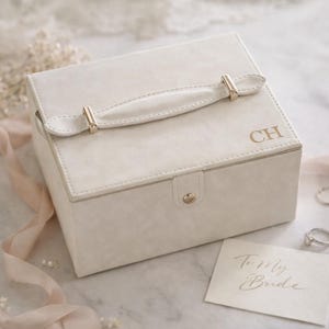Può includere: Portagioie color crema con manico e ferramenta color oro. La scatola ha le iniziali "CH" sulla parte superiore e una carta con la scritta "To My Bride". La scatola è su una superficie di marmo.