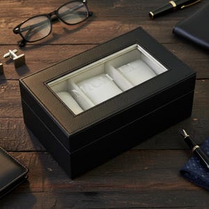 Pu&ograve; includere: Una scatola per orologi in pelle nera con un piano in vetro trasparente, che mostra il nome "MICHAEL" a lettere bianche. La scatola ha tre scomparti con cuscini bianchi. Altri accessori come occhiali, penne e un portafoglio sono su una superficie di legno scuro.