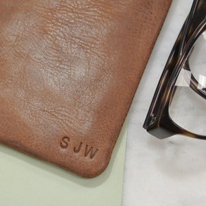 May include: A brown leather pouch with the initials "SJW" embossed on the front. The pouch is lying on a light green surface next to a pair of tortoiseshell glasses.