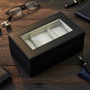 Pu&ograve; includere: Una scatola per orologi in pelle nera con un coperchio in vetro trasparente, che mostra il nome "MICHAEL" a lettere bianche. La scatola ha tre scomparti con cuscini bianchi. Sono visibili accessori come occhiali, gemelli e una penna.