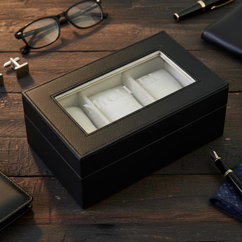 Pu&ograve; includere: Una scatola per orologi in pelle nera con un coperchio in vetro trasparente, che mostra il nome "MICHAEL" a lettere bianche. La scatola ha tre scomparti con cuscini bianchi. Sono visibili accessori come occhiali, gemelli e una penna.