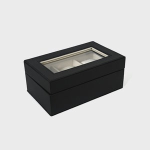Pu&ograve; includere: Una scatola per orologi nera di forma rettangolare. La scatola ha un coperchio in vetro trasparente, che rivela due scomparti con interni bianchi. L'esterno &egrave; di colore nero pieno.