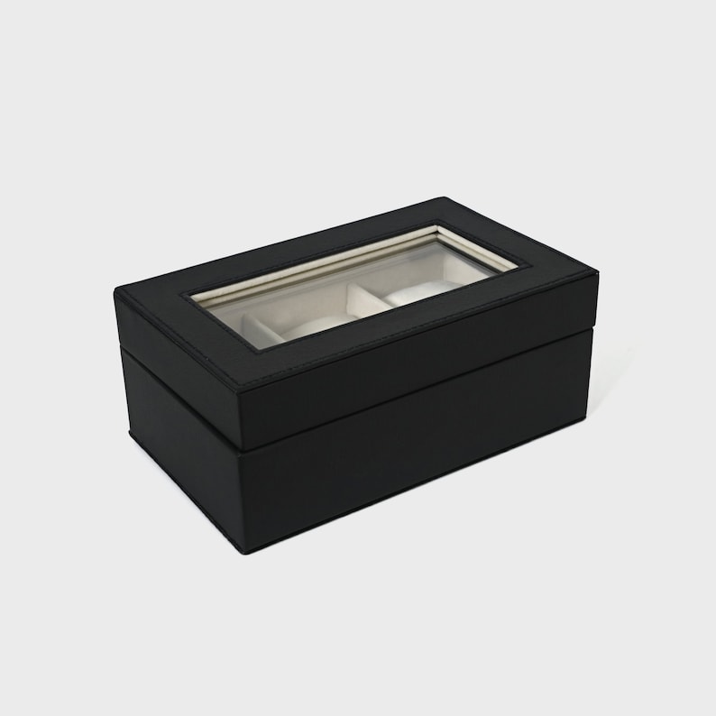 Pu&ograve; includere: Una scatola per orologi nera di forma rettangolare. La scatola ha un coperchio in vetro trasparente, che rivela due scomparti con interni bianchi. L'esterno &egrave; di colore nero pieno.