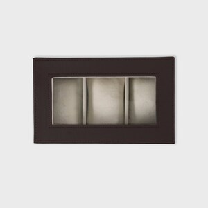Pu&ograve; includere: Una scatola per orologi rettangolare in pelle marrone con una finestra trasparente, che mostra tre scomparti individuali con cuscini colorati chiari. La scatola ha bordi cuciti e un design minimalista, adatta per riporre orologi.