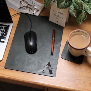 May include: A dark grey mouse mat with the name "Andrew" and the letter "A" engraved. A black computer mouse, a pen, and a cup of tea are on the mat. A "Happy Father's Day" card is also visible.