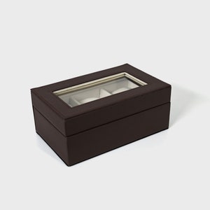 Pu&ograve; includere: Una scatola per orologi rettangolare marrone scuro con un piano in vetro trasparente, che mostra tre cuscini bianchi. La scatola &egrave; realizzata in materiale marrone scuro con cuciture visibili sui bordi. Design semplice ed elegante.