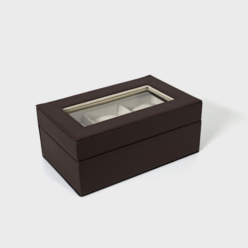 Pu&ograve; includere: Una scatola per orologi rettangolare marrone scuro con un piano in vetro trasparente, che mostra tre cuscini bianchi. La scatola &egrave; realizzata in materiale marrone scuro con cuciture visibili sui bordi. Design semplice ed elegante.