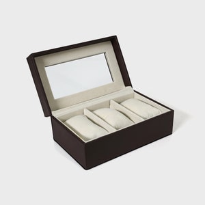 Pu&ograve; includere: Una scatola per orologi marrone con coperchio incernierato e specchio rettangolare all'interno. L'interno presenta tre cuscini per orologi color crema. La scatola &egrave; progettata per riporre e proteggere gli orologi, mostrando un design classico ed elegante.
