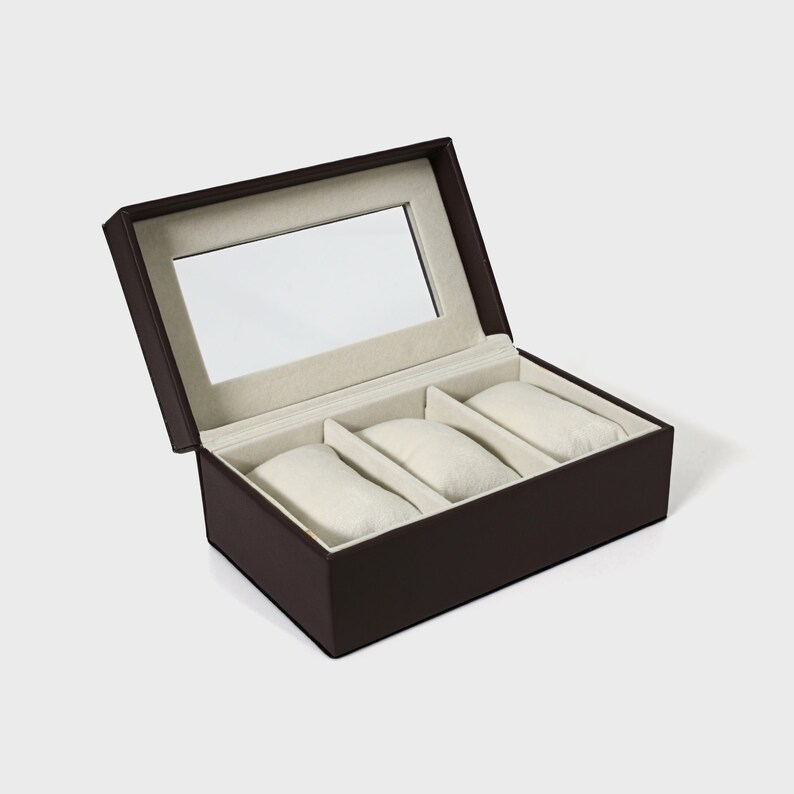 Pu&ograve; includere: Una scatola per orologi marrone con coperchio incernierato e specchio rettangolare all'interno. L'interno presenta tre cuscini per orologi color crema. La scatola &egrave; progettata per riporre e proteggere gli orologi, mostrando un design classico ed elegante.