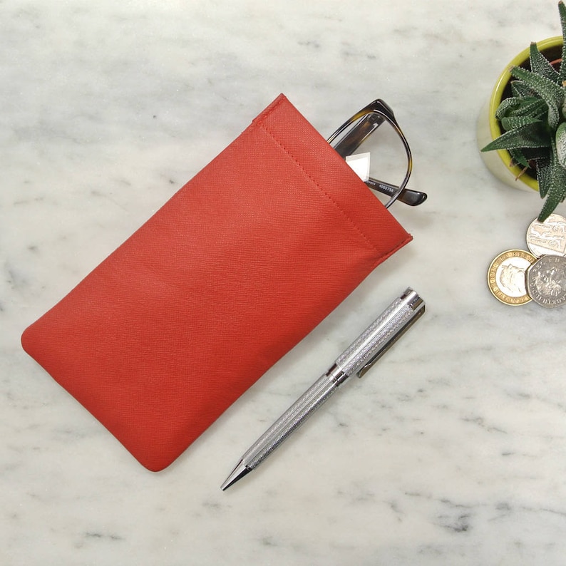 May include: A red fabric glasses case with a pair of glasses inside. A silver pen is lying on a white marble surface.