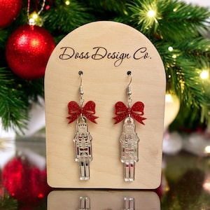 Könnte beinhalten: Silberne Nussknacker-Ohrringe mit roten Glitzerbögen. Die Ohrringe werden auf einem Holzständer mit dem Text "Doss Design Co." präsentiert.