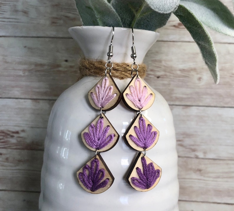 May include: A pair of dangle earrings with three tiers of wooden teardrop shapes. Each teardrop is embroidered with a floral design in shades of pink and purple.