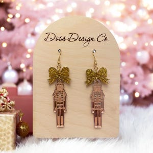 Op de afbeelding: Oorbellen met gouden glitterboogjes en een roze gouden notenkrakerbedel. De oorbellen worden getoond op een houten standaard met de tekst "Doss Design Co."