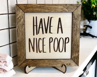 May include: A wooden sign with a brown frame and a white background. The sign says "Have a Nice Poop" in black lettering.