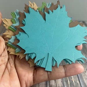 May include: A stack of teal paper maple leaves with brown and green paper leaves underneath.