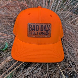 May include: Bright orange trucker hat with a curved brim and mesh back. The hat features a brown leather patch with the text "BAD DAY TO BE A SPIKE" and a deer silhouette.