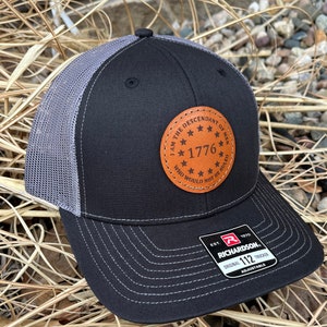 May include: Black trucker hat with a gray mesh back and a curved brim. The hat features a brown leather patch with the text "I AM THE DESCENDANT OF MEN WHO WOULD NOT BE RULED 1776". The hat is adjustable.