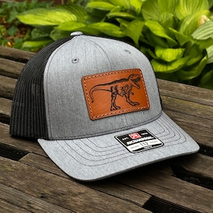 May include: A gray and black trucker hat with a brown leather patch featuring a black silhouette of a Tyrannosaurus Rex. The hat has a black mesh back and a gray front with a black stitched brim. The hat has a black label with the text "EST. 1913 RICHARDSON 112 ORIGINAL TRUCKER YOUTH".