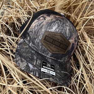May include: A camouflage trucker hat with a black mesh back. The hat has a brown leather patch that says "Custom Patch Your Logo Here! Shapes Available". The hat also has a brown leather label that says "Est. 1970 Richardson. Original 112 Trucker Adjustable".