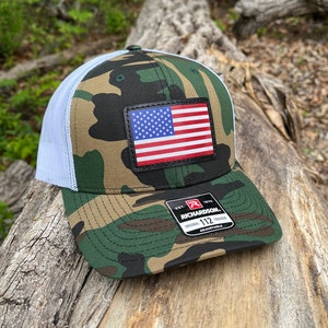 May include: Camouflage pattern trucker hat with a white mesh back and a leather patch featuring the American flag. The hat has a curved brim and an adjustable strap. The brand name "Richardson" is visible on a tag.