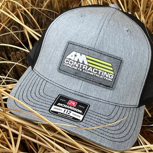 May include: A gray and black trucker hat with a black mesh back. The hat has a patch on the front that says "AM Contracting Concrete Construction". The hat is made by Richardson 112.