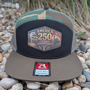 May include: A snapback cap with a flat bill, featuring a camouflage pattern on the crown and a solid olive-drab brim. A hexagonal patch on the front displays the text AMERICA 250 YEARS, 1776-2026, and imagery of an eagle, Mount Rushmore, and a flag.