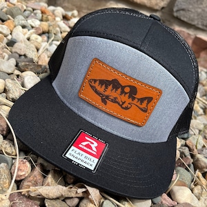 May include: A black and grey baseball cap with a brown leather patch featuring a fish silhouette. The cap has a mesh back and a flat bill. The label on the cap reads "Richardson Flat Bill Snapback Est 1910."