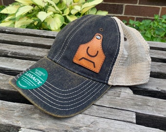 May include: A black and tan trucker hat with a brown leather patch featuring a stylized letter "C". The hat has a mesh back and a distressed look. The hat has a sticker that says "Old Favorite Trucker" and "Legacy".