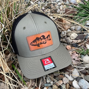May include: A green and black trucker hat with a brown leather patch featuring a bass fish silhouette. The patch has a forest scene with a full moon. The hat has a black mesh back and a red label that says "Richardson" and "Flexfit".
