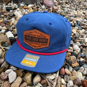 May include: A blue and red baseball cap with a brown leather patch that says "Custom Patch Your Logo Here! Shapes Available". The cap is on a bed of small rocks.