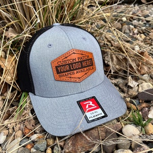 May include: A gray and black baseball cap with a brown leather patch that says "CUSTOM PATCH YOUR LOGO HERE! SHAPES AVAILABLE". The cap has a black mesh back and a gray front. The cap has a Richardson Flexfit label on the side.