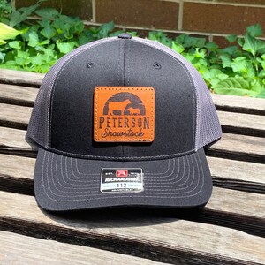 May include: A black and gray trucker hat with a brown leather patch that says "Peterson Showstock" with a silhouette of two cows. The hat is adjustable and has a Richardson label.