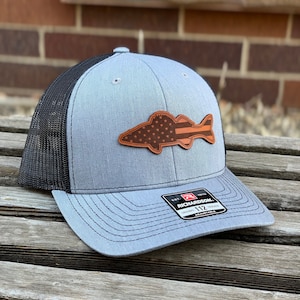 May include: A gray and black trucker hat with a brown leather patch of a fish with an American flag design. The patch has a red stripe across the fish. The hat has a black mesh back and a gray front. The hat has a Richardson 112 adjustable tag.