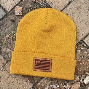 May include: A mustard yellow knit beanie with a brown leather patch featuring a silhouette of a cow and an American flag.