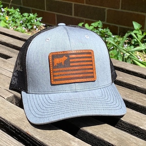 May include: A gray trucker hat with a black mesh back and a curved brim. The hat features a brown leather patch with a cow silhouette and American flag design. The hat is sitting on a wooden surface.