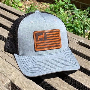 May include: A gray trucker hat with a black mesh back. The front features a brown leather patch with an American flag design and a llama silhouette. The hat is sitting on a wooden surface.