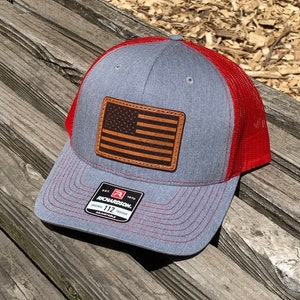 May include: A gray and red trucker hat with a brown leather American flag patch. The hat is adjustable and has the Richardson logo on the side.