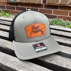 May include: A green and black trucker hat with a brown leather patch that says "Keep Choppin'" in cursive. The hat is adjustable and has a black mesh back.