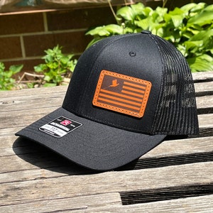 May include: Black trucker hat with a mesh back and a curved brim. The hat features a rectangular patch with an orange and brown flag design. The patch has a bird graphic. The hat is adjustable.