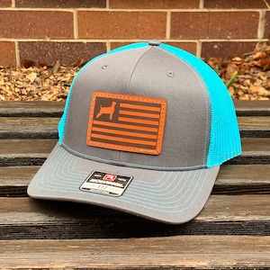 May include: A gray trucker hat with a turquoise mesh back and brim. The front features a brown leather patch with an orange flag design and a dog silhouette. The hat has a curved brim and a snapback closure.