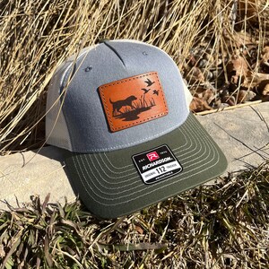 May include: A gray and green trucker hat with a brown leather patch featuring a dog and ducks. The patch says "Richardson 112 Adjustable".