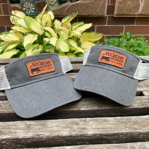 May include: Two gray visors with white mesh sides and a brown leather patch. The patch features the text "McCreary Gelbvieh Est. 1979" and a silhouette of a cow. The visors are displayed on a wooden surface.