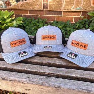 May include: Three gray trucker hats with black mesh backs and light blue brims are displayed. Each hat has a rectangular, orange leather patch with a name: "WHITING", "SIMPSON", and "OWENS". The hats are on a wooden bench.