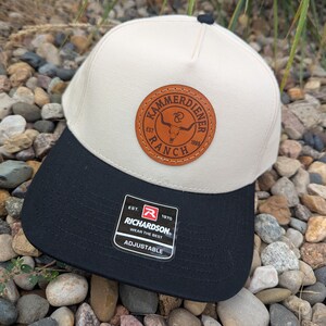 May include: A beige and black baseball cap with a leather patch that reads "KAMMERDIENER RANCH" and the Richardson logo. The cap has an adjustable strap.