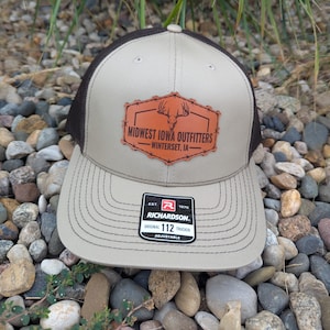 May include: A tan and brown trucker hat with a curved brim. The hat features a leather patch with the text "Midwest Iowa Outfitters" and a deer head logo. The hat also has a Richardson 112 tag.