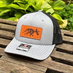 May include: A gray and black trucker hat with a brown leather patch featuring a triceratops skeleton. The patch has stitching around the edges. The hat has a black mesh back and a gray front with a black Richardson logo.